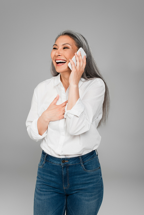 mature woman touching chest and laughing while talking on mobile phone isolated on grey