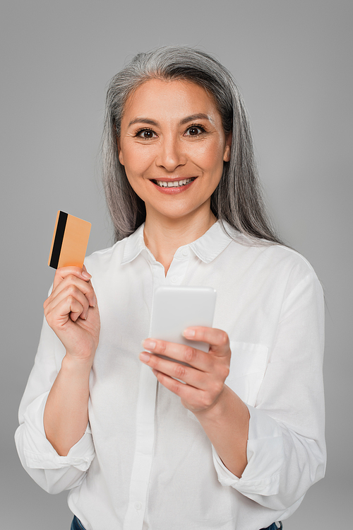 stylish middle aged woman with credit card and mobile phone smiling at camera isolated on grey