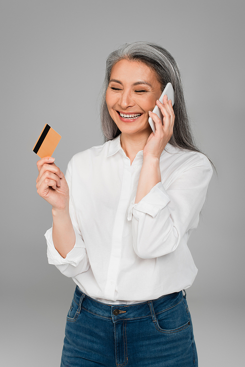 cheerful middle aged woman with credit card talking on cellphone isolated on grey