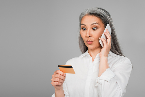 surprised mature woman calling on mobile phone while looking at credit card isolated on grey