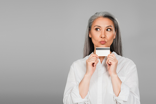 thoughtful mature woman in white shirt holding credit card isolated on grey