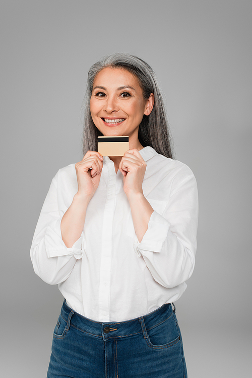 pleased woman in white shirt showing credit card while  isolated on grey
