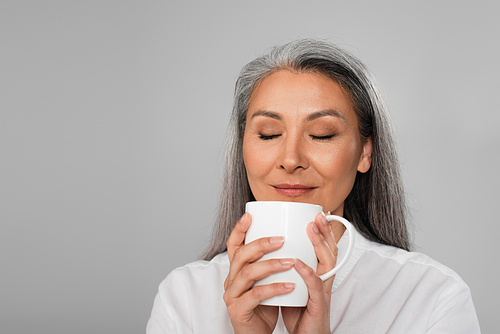 pleased middle aged woman enjoying flavor of tea isolated on grey