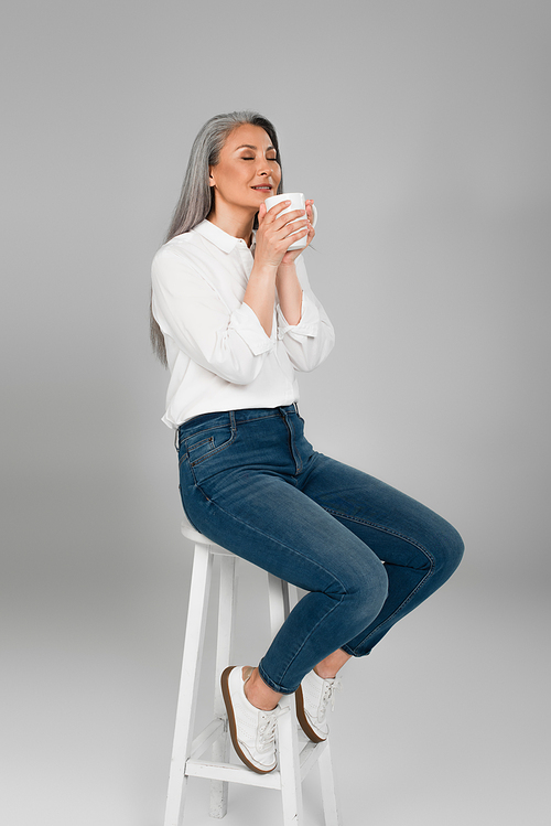 middle aged woman in white shirt and jeans sitting on stool with cup of aromatic tea on grey