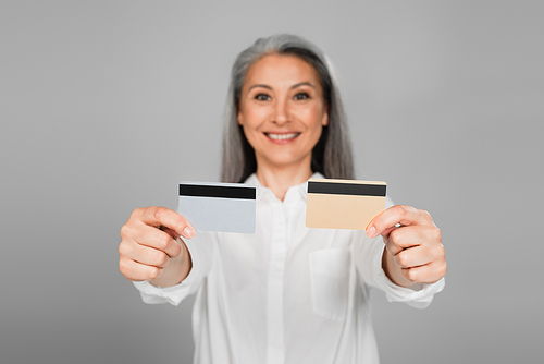 blurred mature woman smiling while showing credit cards isolated on grey