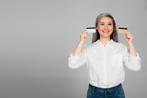 joyful middle aged woman in white shirt showing credit cards isolated on grey