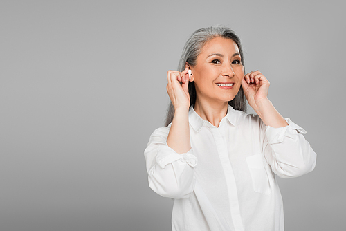 joyful woman with grey hair inserting wireless earphones isolated on grey