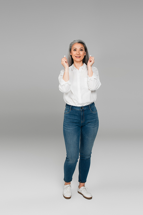 full length view of mature woman in jeans and shirt holding earphones on grey