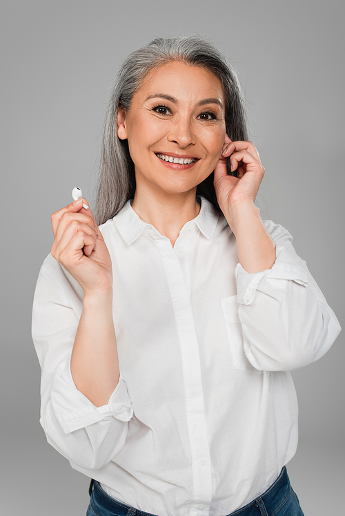 happy middle aged woman in white shirt holding wireless earphone isolated on grey