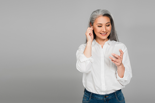 joyful mature woman adjusting earphone while using smartphone isolated on grey