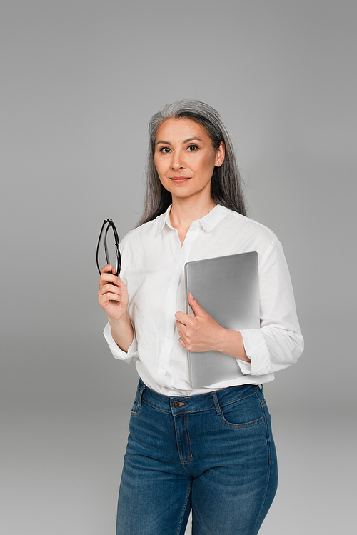 mature woman in white shirt holding eyeglasses and laptop isolated on grey