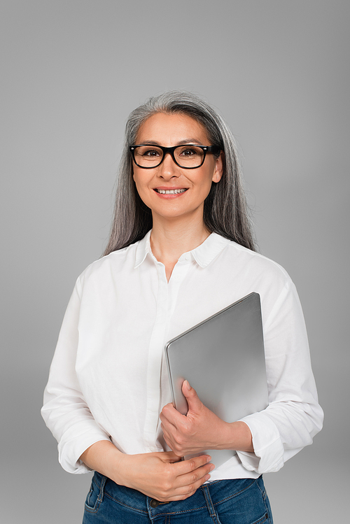middle aged woman in trendy eyeglasses holding laptop and smiling at camera isolated on grey