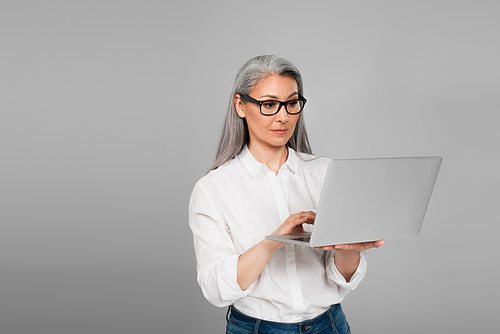 mature woman in shirt and eyeglasses using laptop isolated on grey
