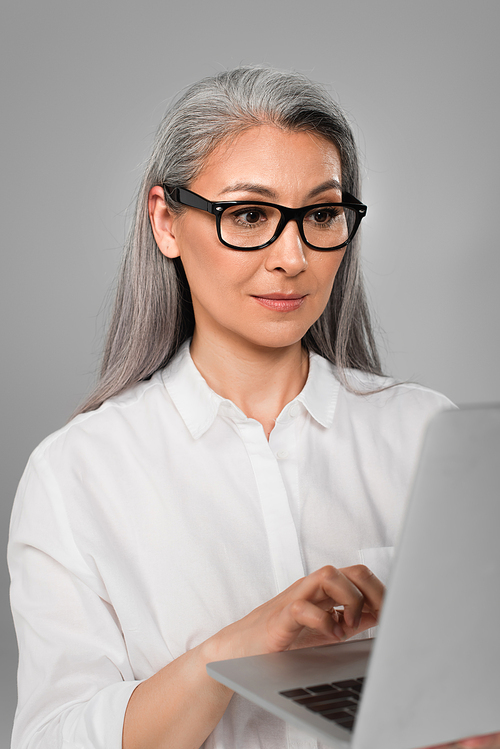 mature woman in white shirt and stylish eyeglasses typing on laptop isolated on grey