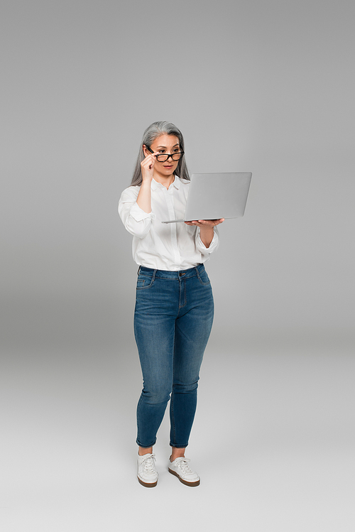 full length view of middle aged woman adjusting eyeglasses while looking at laptop on grey background
