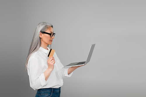 middle aged woman in white shirt and eyeglasses holding laptop and credit card isolated on grey
