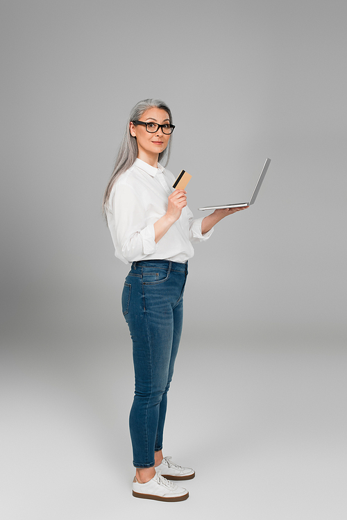 mature woman in jeans and white shirt standing with credit card and laptop on grey background