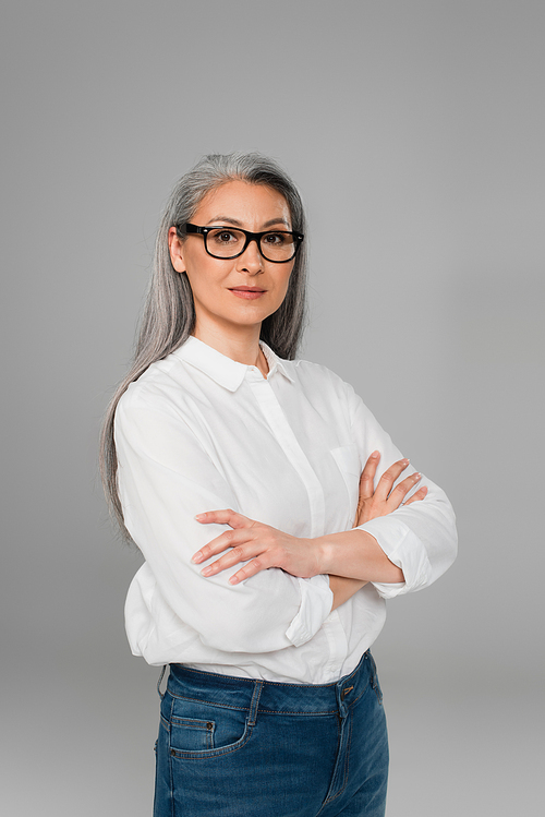 confident mature woman in stylish eyeglasses and white shirt standing with crossed arms isolated on grey
