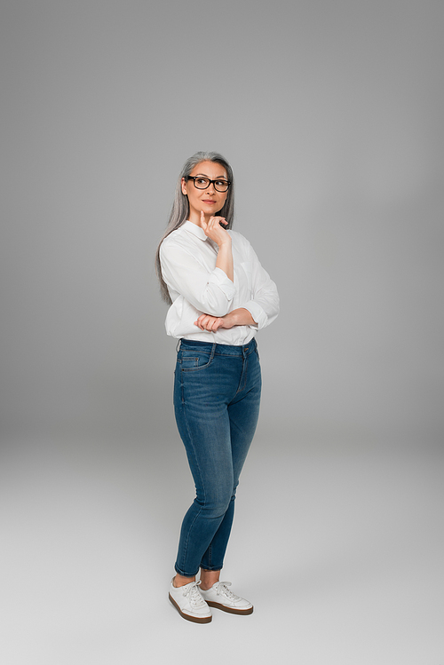 full length view of mature woman in jeans, white shirt and eyeglasses standing on grey