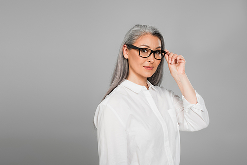 middle aged woman in white shirt  while touching eyeglasses isolated on grey