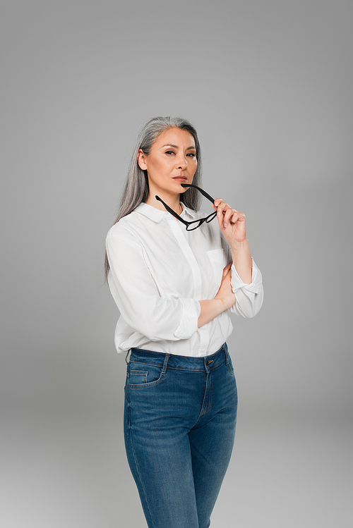mature, confident woman in jeans and white shirt holding eyeglasses isolated on grey