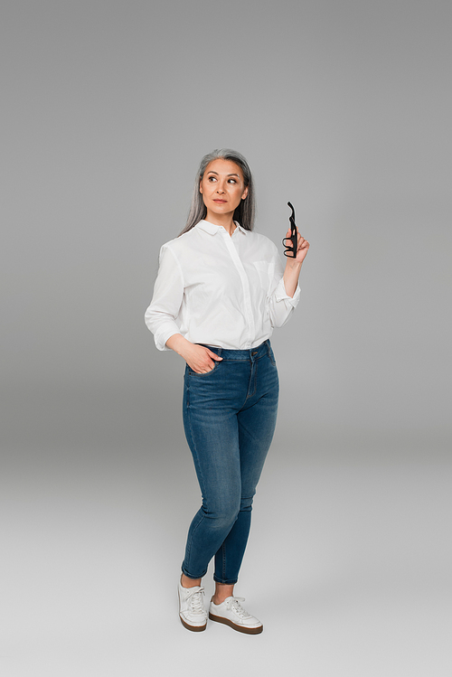 middle aged woman holding eyeglasses while standing with hand in pocket of jeans on grey