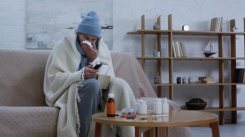 sick man in warm beanie and hat clicking tv channels while suffering from runny nose near medication on table