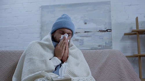 sick man in warm beanie and blanket sneezing in paper napkin while sitting on sofa