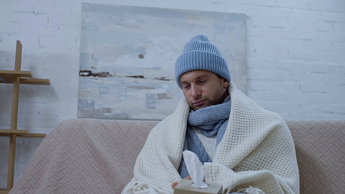 sick man with pack of paper napkins sitting on sofa in warm beanie, scarf and blanket