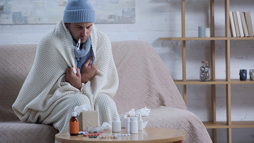 sick man measuring temperature while sitting on couch in warm blanket and beanie near medicaments
