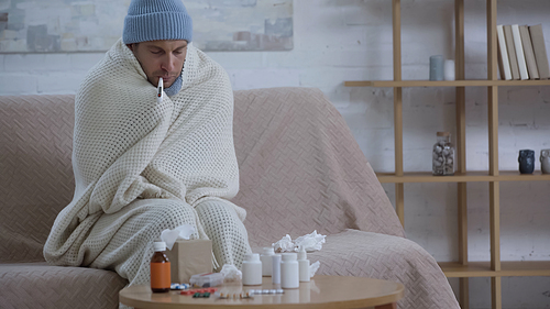 sick man in warm hat and beanie measuring temperature near table with medicaments