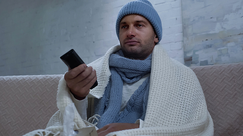 sick man in warm beanie, scarf and blanket clicking channels while watching tv