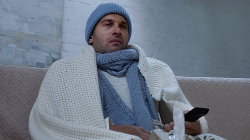 diseased man in warm hat, scarf and blanket watching tv on couch at home