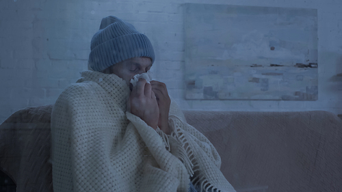sick man sitting on couch in warm beanie and blanket and sneezing in paper napkin