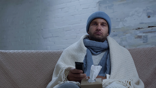 ill man with tv remote controller and paper napkins sitting on couch in warm beanie and blanket