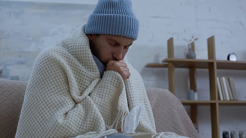 ill man in warm hat and blanket coughing on couch at home