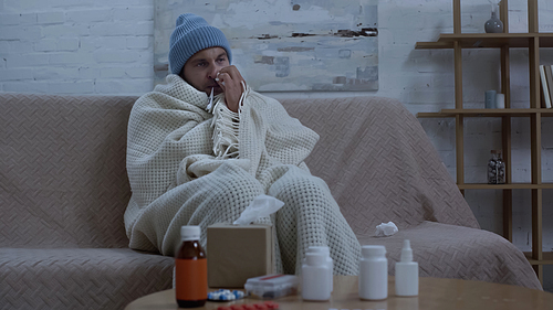diseased man wiping nose with paper napkin while sitting on couch in warm hat and blanket near table with medication