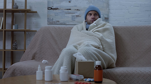 ill man measuring temperature while sitting on couch in warm beanie and blanket near table with medication