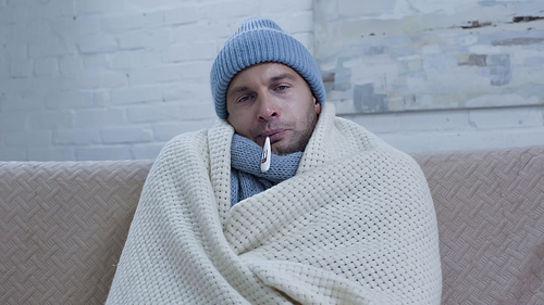 diseased man measuring temperature while sitting on couch in warm beanie and blanket
