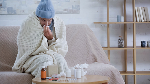 ill man wiping nose with paper napkin while sitting in warm blanket and hat near medicaments on table