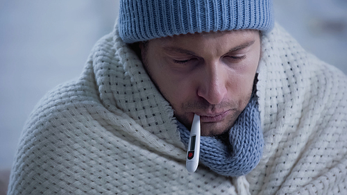ill man in warm blanket and beanie measuring temperature with thermometer in mouth
