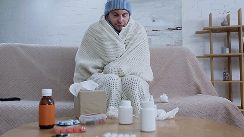 diseased man in warm hat and blanket sitting on couch near table with medication