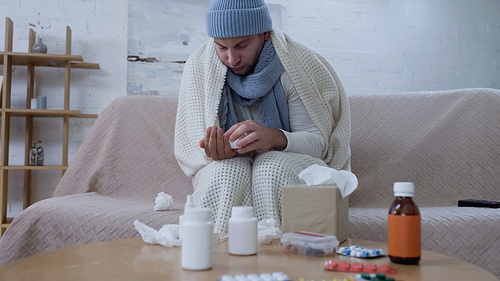 sick man in warm blanket and hat holding pills container near table with medicaments