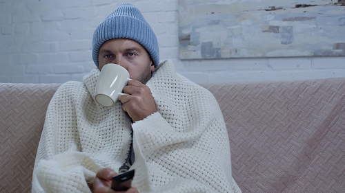 sick man drinking warm beverage and watching tv while sitting on sofa in beanie and blanket