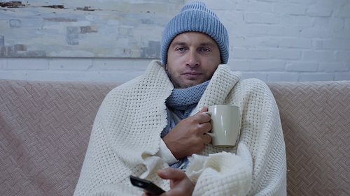 sick man in warm blanket and hat sitting on couch with cup of tea and watching tv