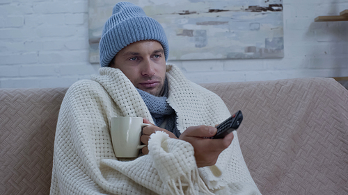 ill man with cup of tea and tv remote controller sitting on couch in warm hat and blanket