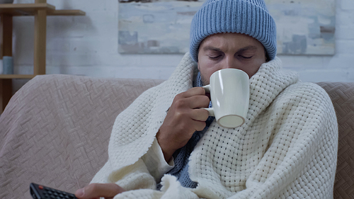diseased man with tv remote controller drinking tea while sitting on couch in warm beanie and blanket