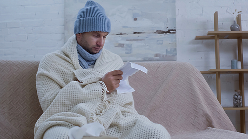ill man in warm beanie and blanket reading medication instruction on couch at home