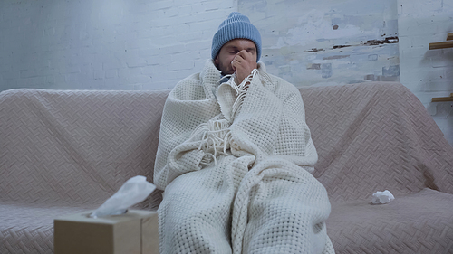 sick man sneezing in paper napkin while sitting on sofa in warm beanie and blanket