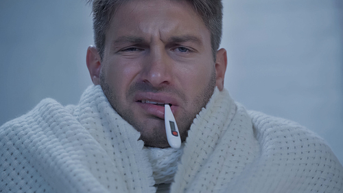 displeased sick man with thermometer in mouth measuring temperature at home